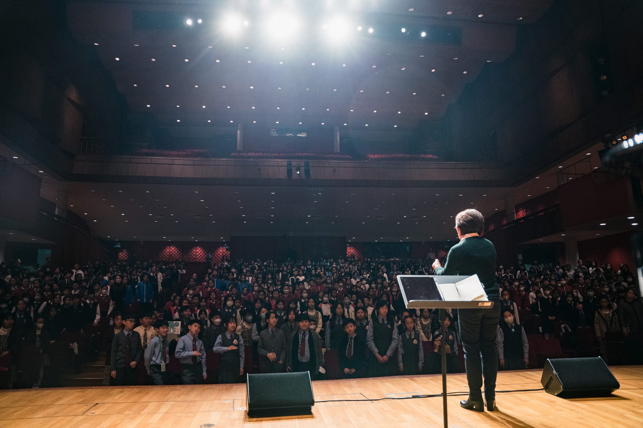A conductor on stage facing a concert hall filled with schoolchildren